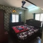 Traditional bedroom with wooden king-size bed, floral bedsheet, ceiling fan, natural lighting, and wardrobe – ideal for homestay, vacation rental, or furnished apartment.