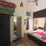A cozy bedroom with a double bed covered in beige and pink bedding and a colorful red and green blanket placed on top. The room has tiled flooring and cream-colored walls. Two barred windows with brown patterned curtains allow natural light in. A ceiling fan and a decorative hanging light fixture are visible. Above the bed is a set of dark wooden overhead cabinets. To the left, a wardrobe with folded mattresses on top stands near a door that leads to a small attached bathroom, which contains a blue bucket. The room is neatly arranged and decorated with framed pictures on the walls.